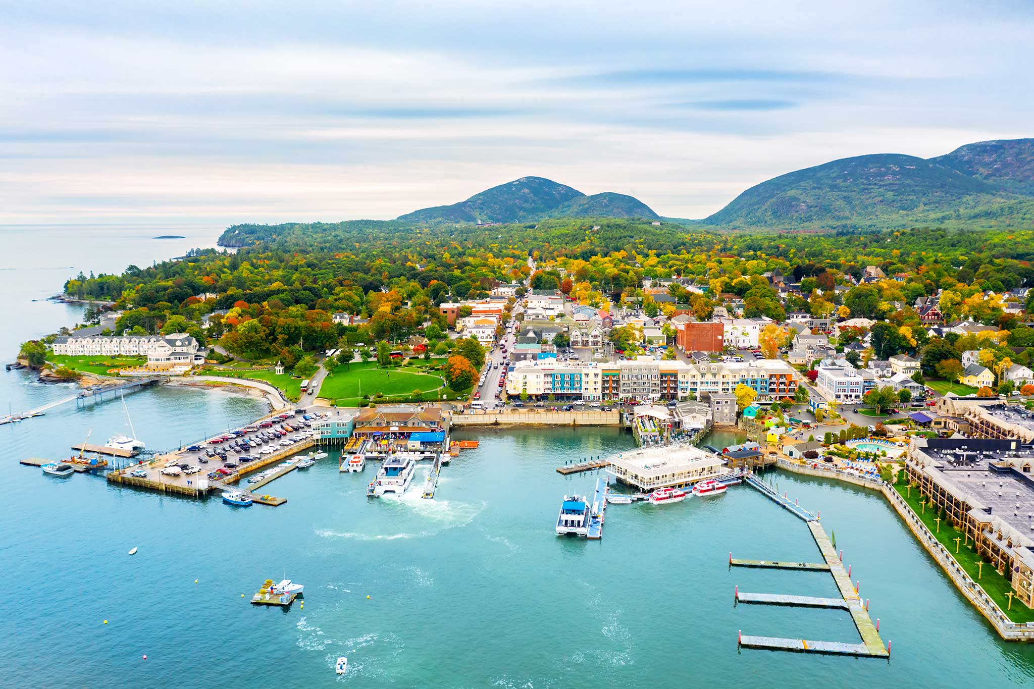 Panoramic view above a quaint seaside town with a marina full of boats, set against a backdrop of lush green hills and a tapestry of fall foliage under an overcast sky.