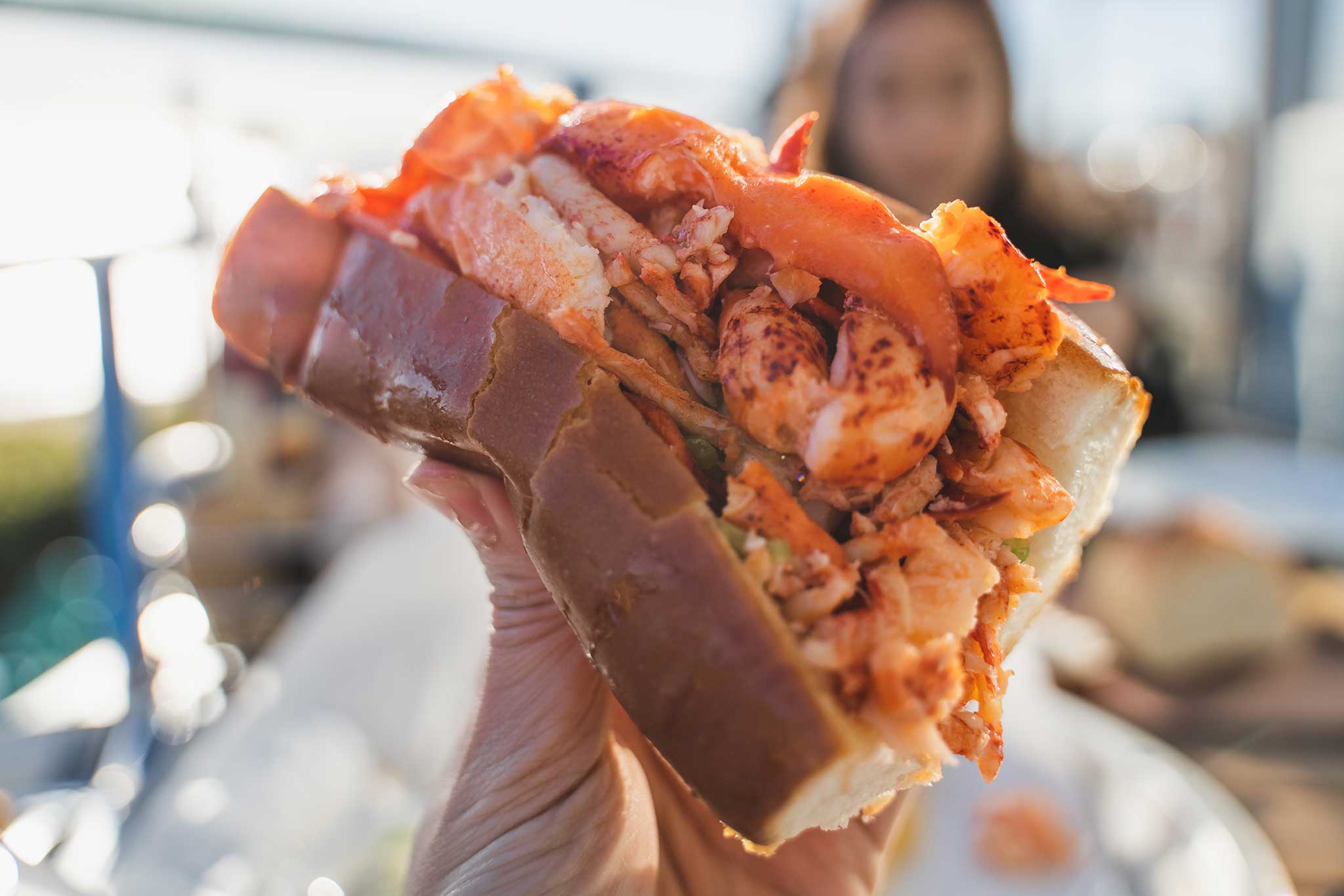 A freshly prepared lobster roll with generous servings of succulent lobster meat held up close for the camera on a beautiful, sunny day.