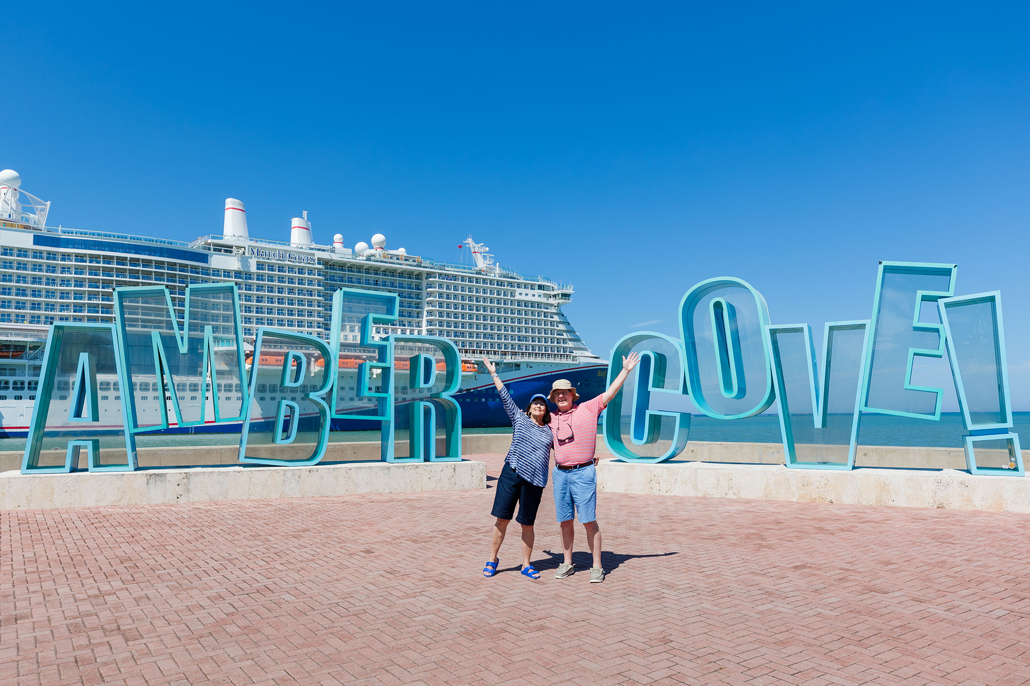 A joyful middle-aged couple posing with raised arms in front of a large “Amber Cove” sign, with a massive cruise ship docked in the background under a clear blue sky.