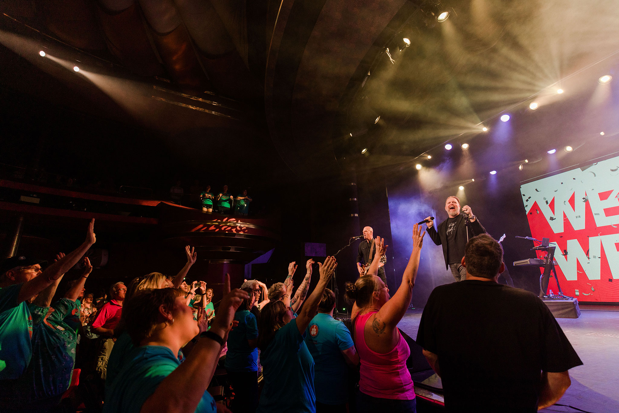 An engaging moment at a concert with a singer onstage and enthusiastic fans in the foreground, hands uplifted, in a dimly lit theater with colorful stage lighting.