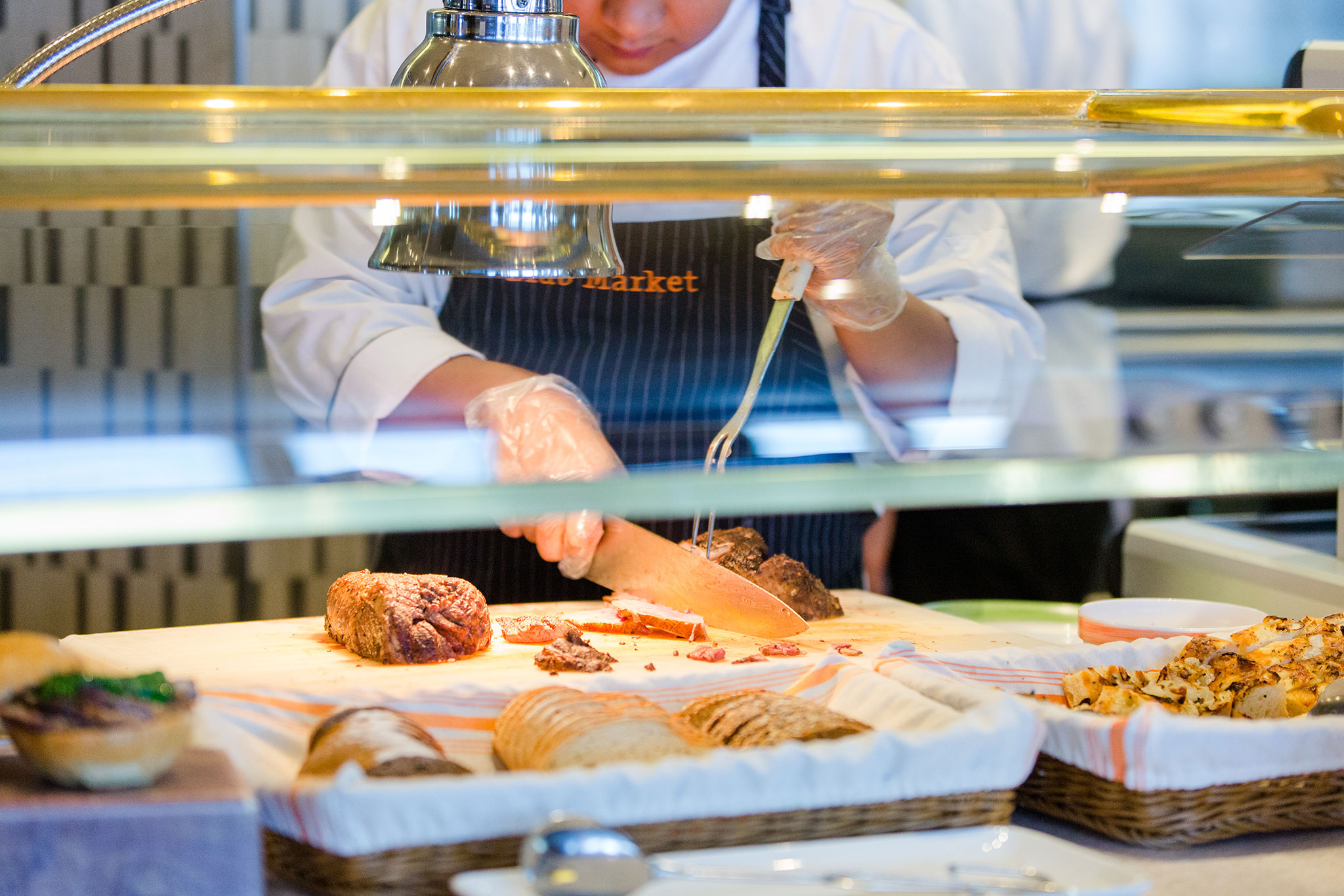 A chef in a striped apron slicing roasted meat at a buffet station, with assorted breads and dishes visible under a bright light.