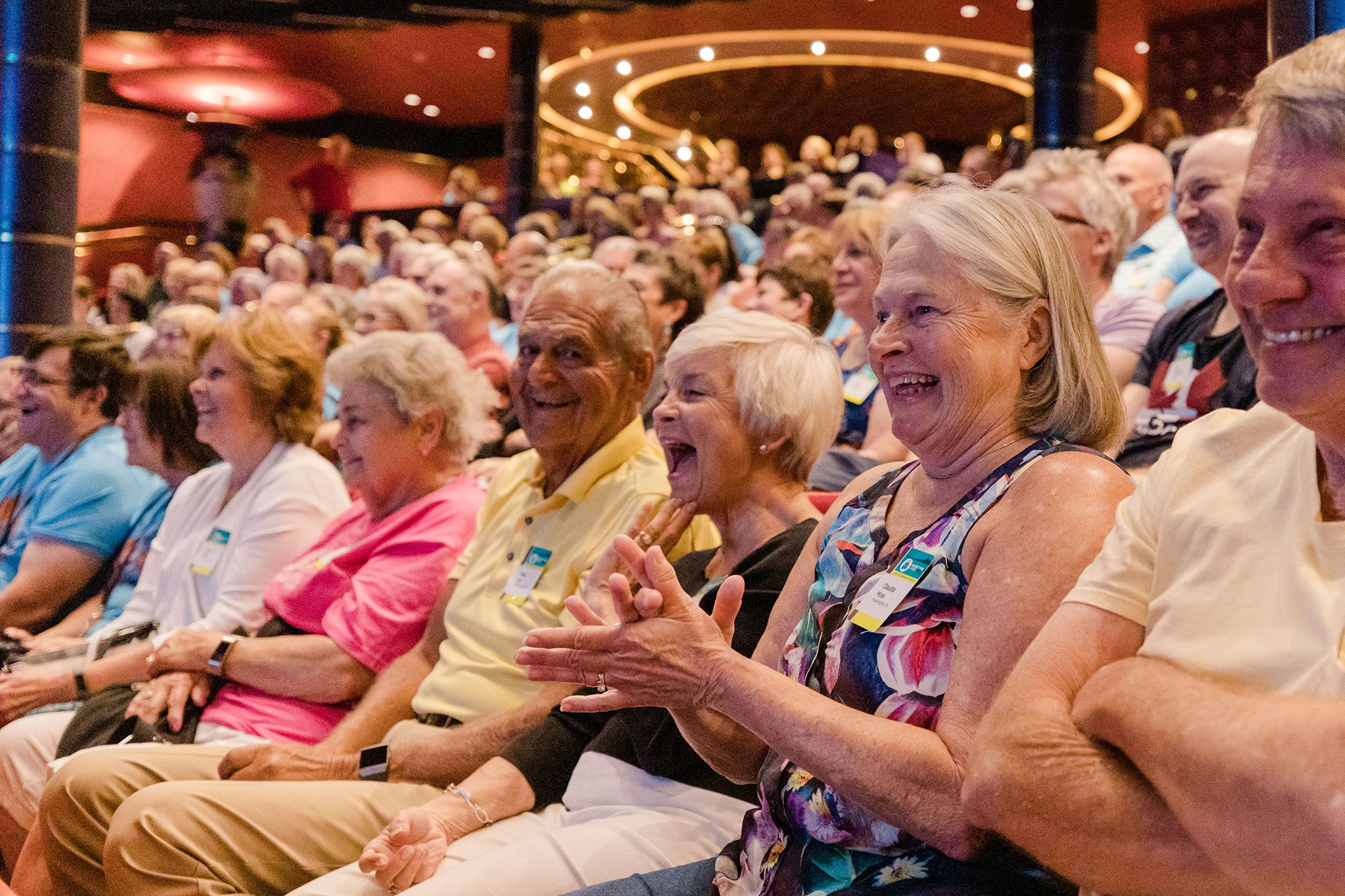 A group of elderly audience members enjoying a show in a theater, with many of them laughing and clapping, wearing colorful casual clothes and name tags.