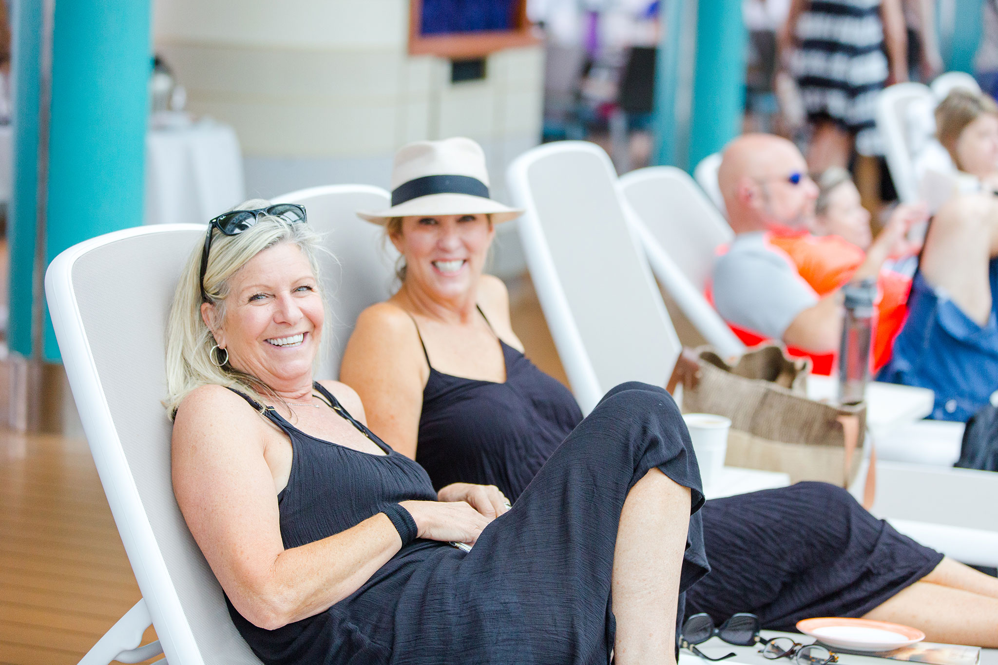 Two women smiling and relaxing on lounge chairs by a poolside; one wearing a black dress and sunglasses on her head, the other in a black dress with a white hat.