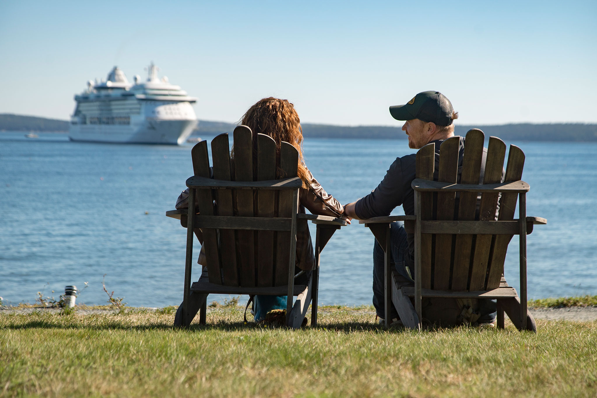 A serene moment captured as a couple, seated on classic wooden chairs, overlooks a tranquil sea with a cruise ship in the distance, emphasizing a peaceful maritime scene.