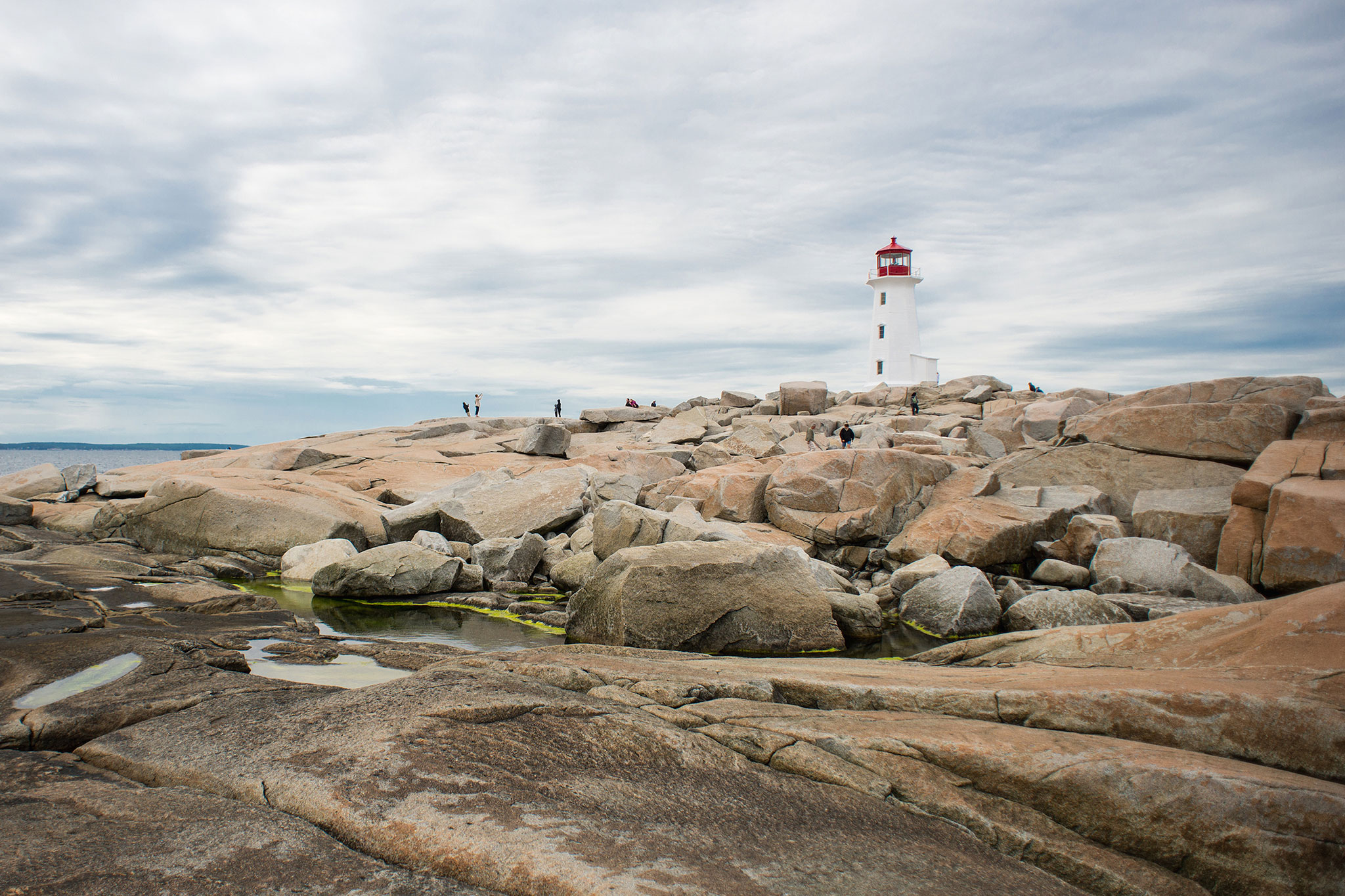 Peggy's Cove Lighthouse stands atop a rugged granite rock landscape under a cloudy sky, with visitors scattered across the rocks exploring the scenic area.