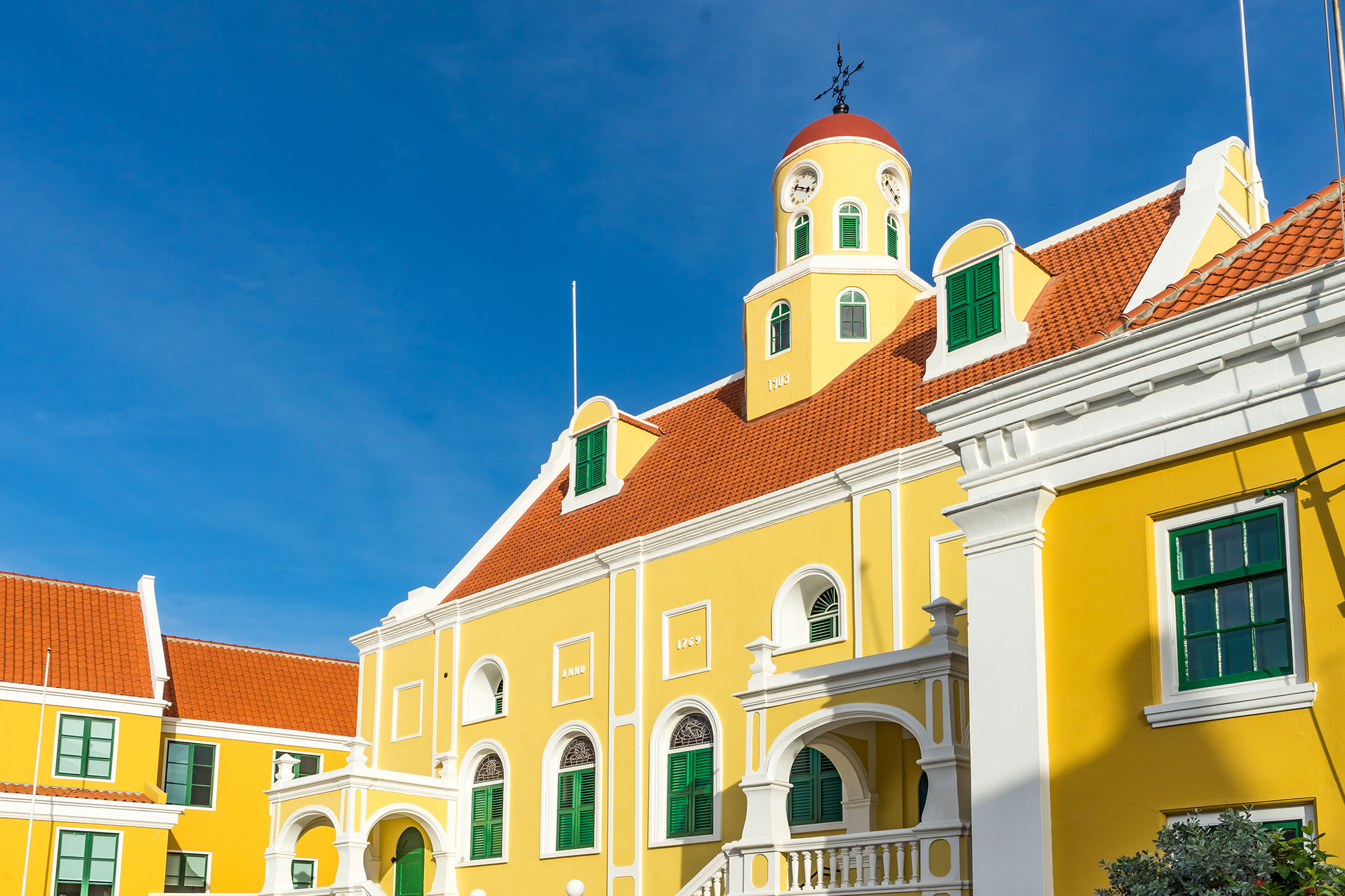 Bright yellow colonial-style building with white trim and red-tiled roofs under a clear blue sky, featuring Dutch arched windows and a clock tower.