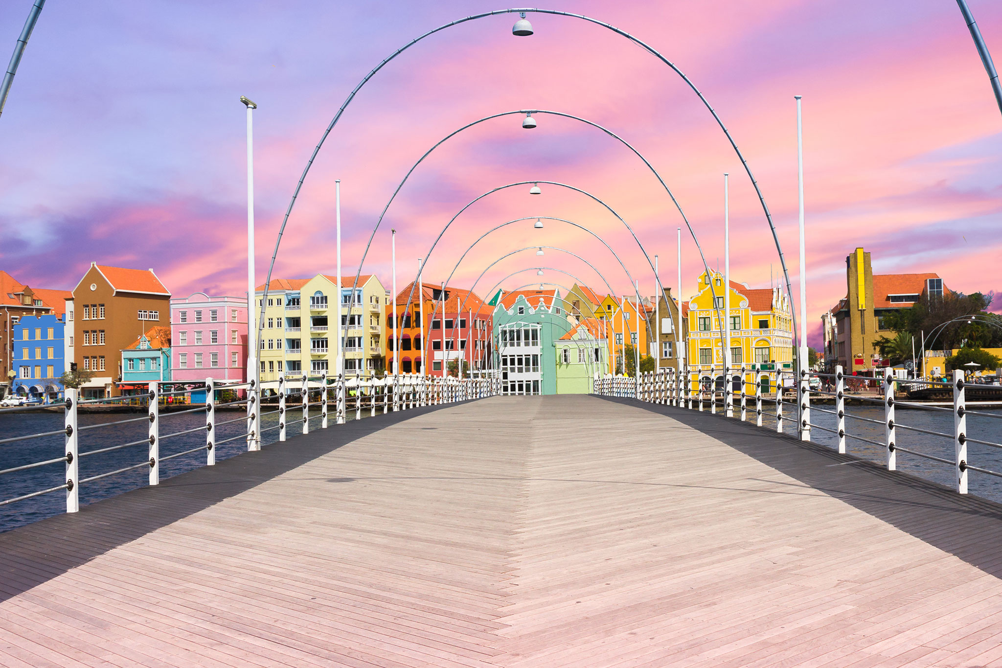 A panoramic view of the Queen Emma Bridge featuring a wooden walkway adorned with arched lights leading toward a row of colorful Dutch colonial-style buildings under a vibrant pink sunset sky.