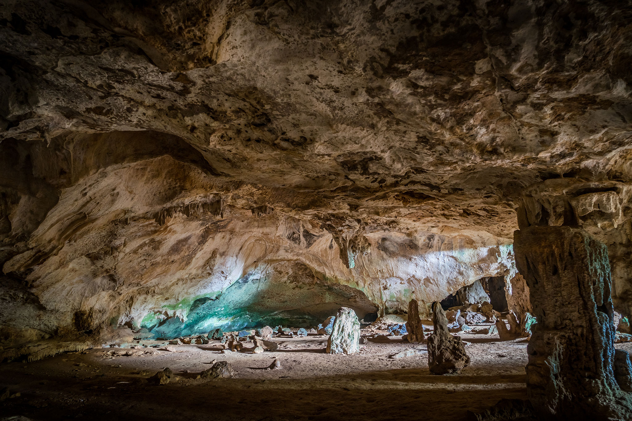 Mysterious interior of a spacious cave with rough textured walls and ceiling, illuminated by subtle lighting that highlights the formations and the rocky ground.