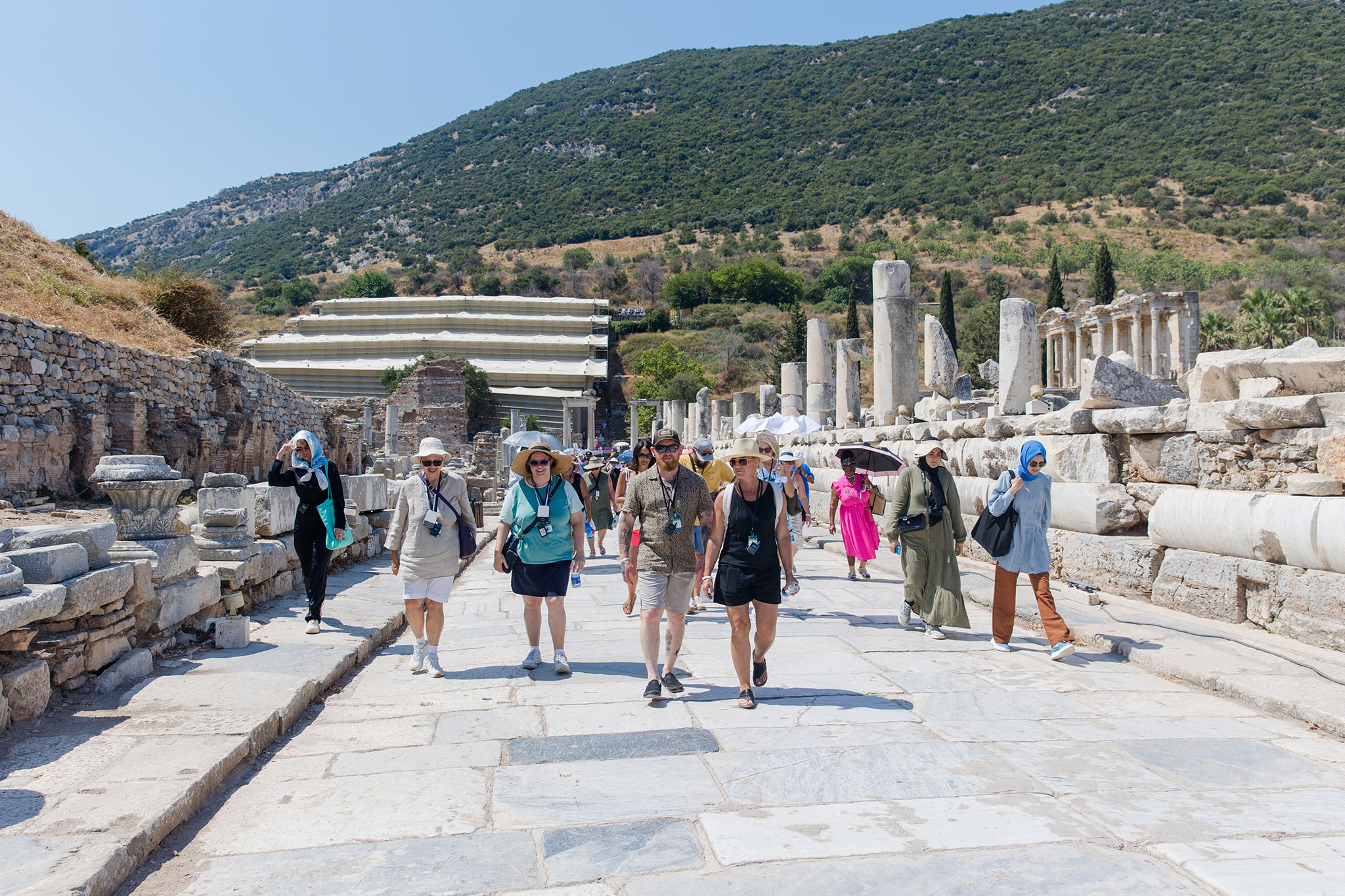 Christian travelers walking along a stone-paved road among ancient ruins in Ephesus, with lush hills in the background.