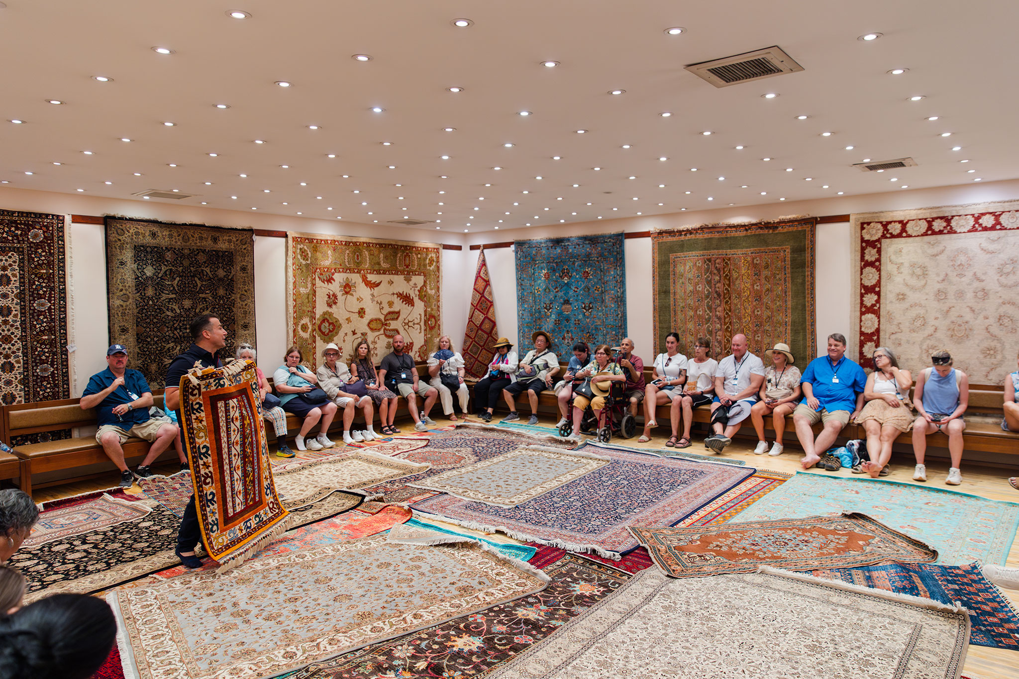 Travelers watching a carpet presentation in a showroom adorned with various traditional carpets.