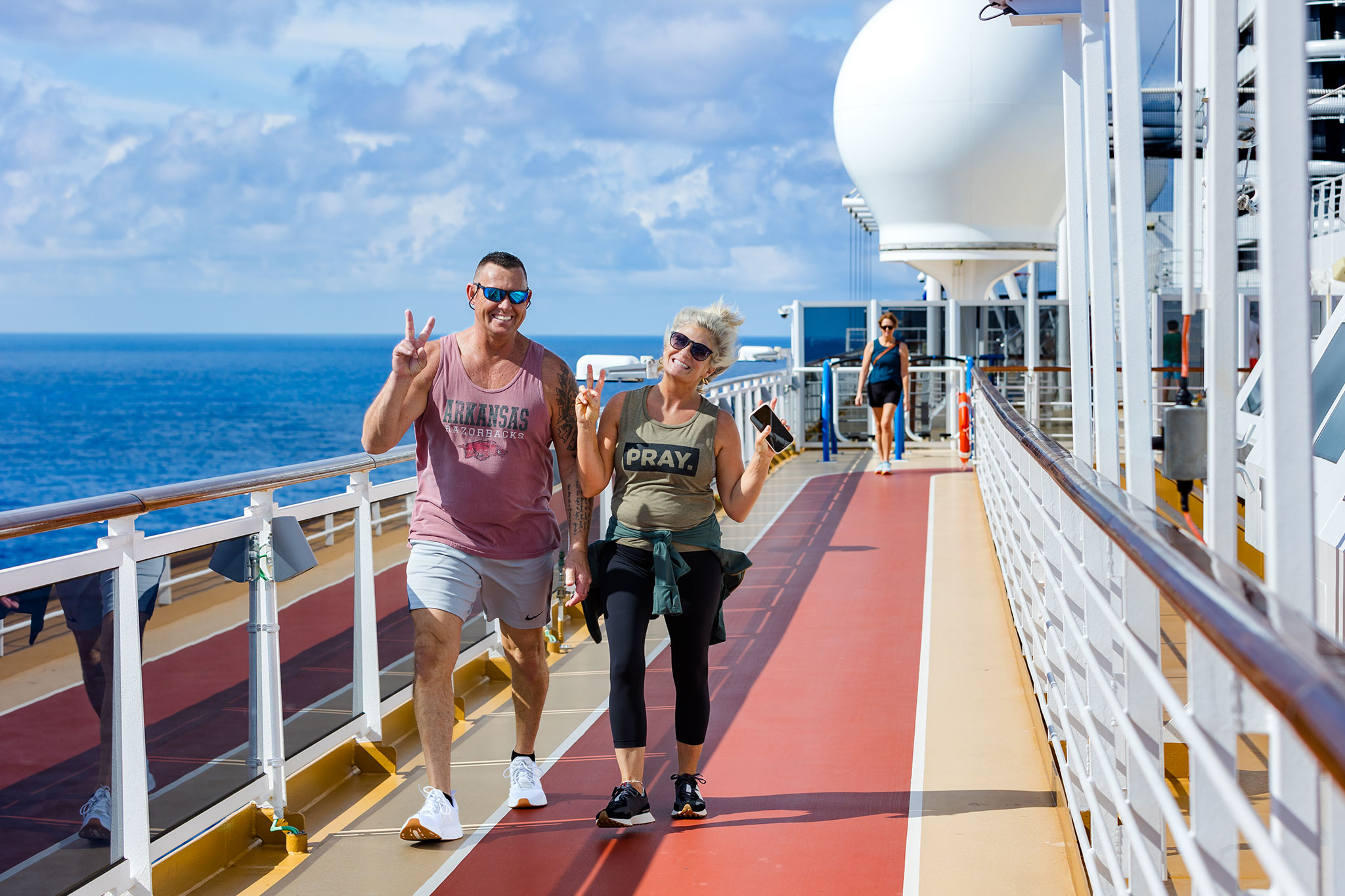 A couple walks on a cruise ship track, smiling and making peace signs, with the ocean and clear skies in the background.