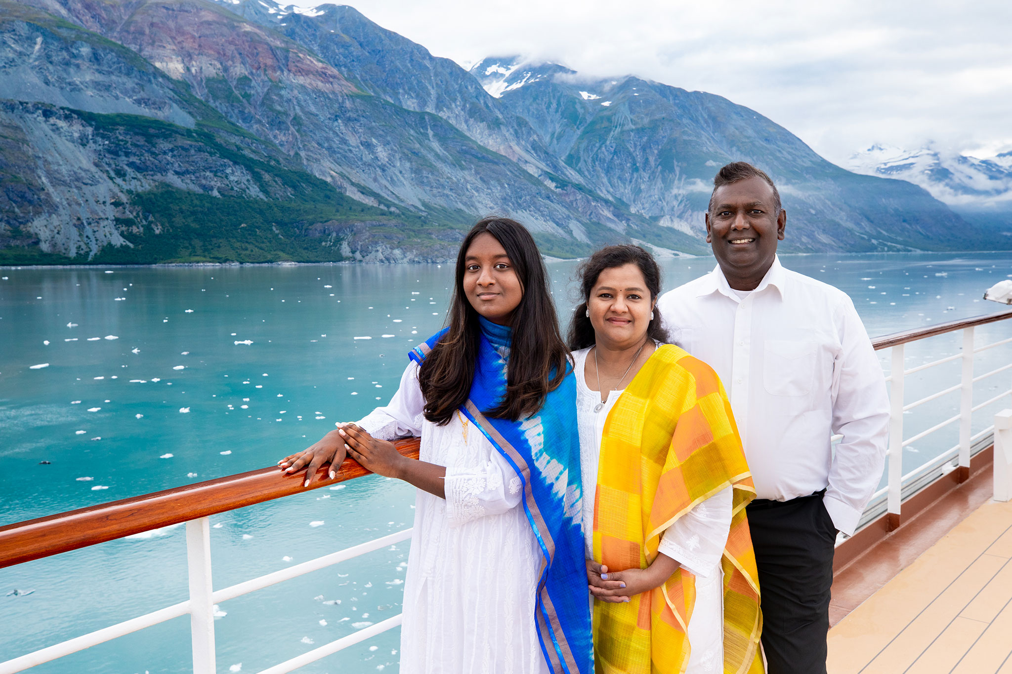 A smiling Indian family, dressed in traditional attire, enjoying a scenic view of a glacier bay from the deck of a cruise ship.