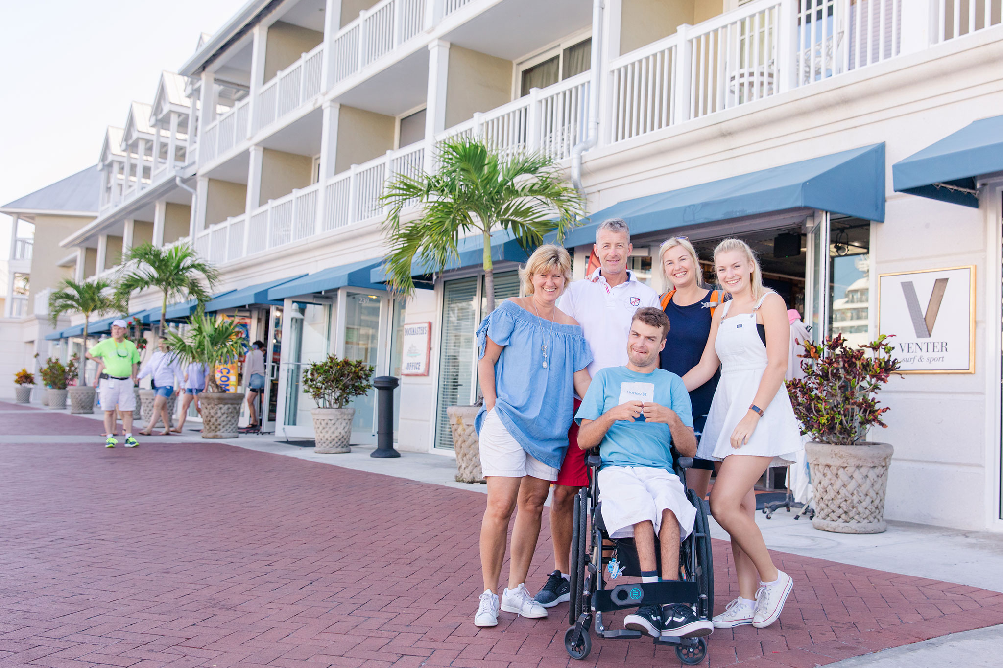 A multigenerational family, including a man in a wheelchair, posing happily together on a sunny sidewalk in front of a row of shops with balconies.