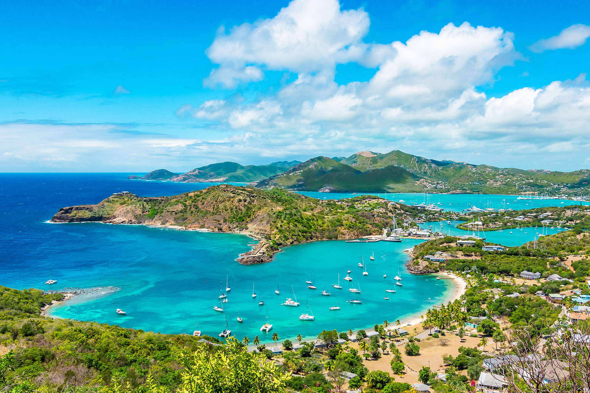 Panoramic view of a tropical bay with turquoise waters, scattered boats, and surrounding lush greenery, under a vibrant blue sky with fluffy clouds.