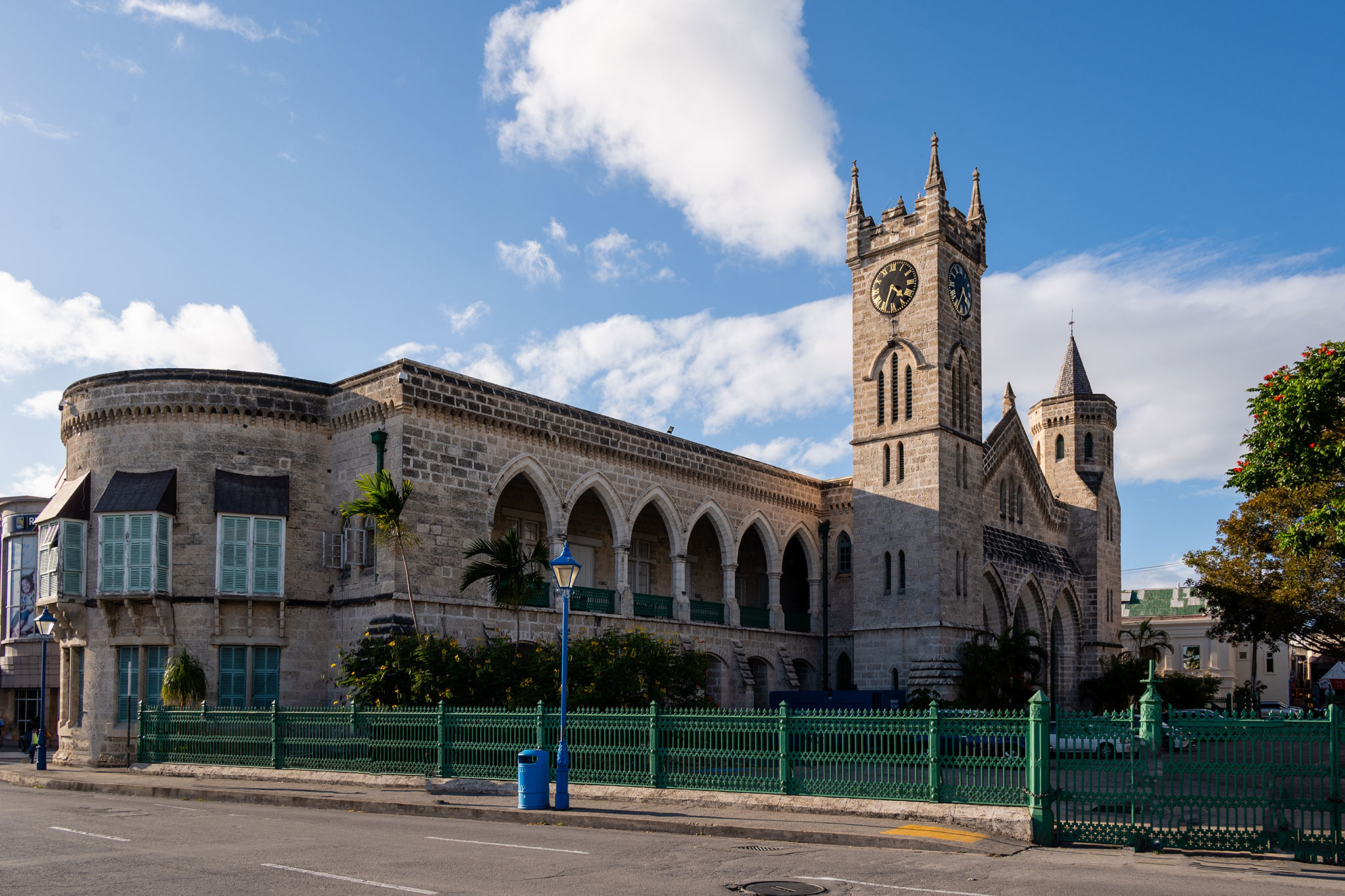 A stone-built Gothic-style church with a clock tower, located on a city street lined with a green fence under a clear blue sky.