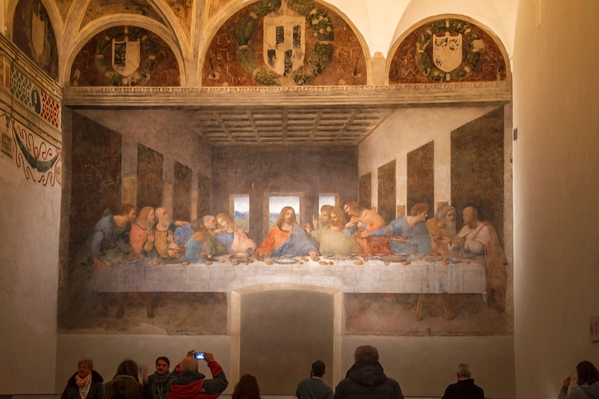 Visitors observing Leonardo da Vinci's The Last Supper mural in a dimly lit room, showcasing the iconic scene of Jesus dining with his disciples.