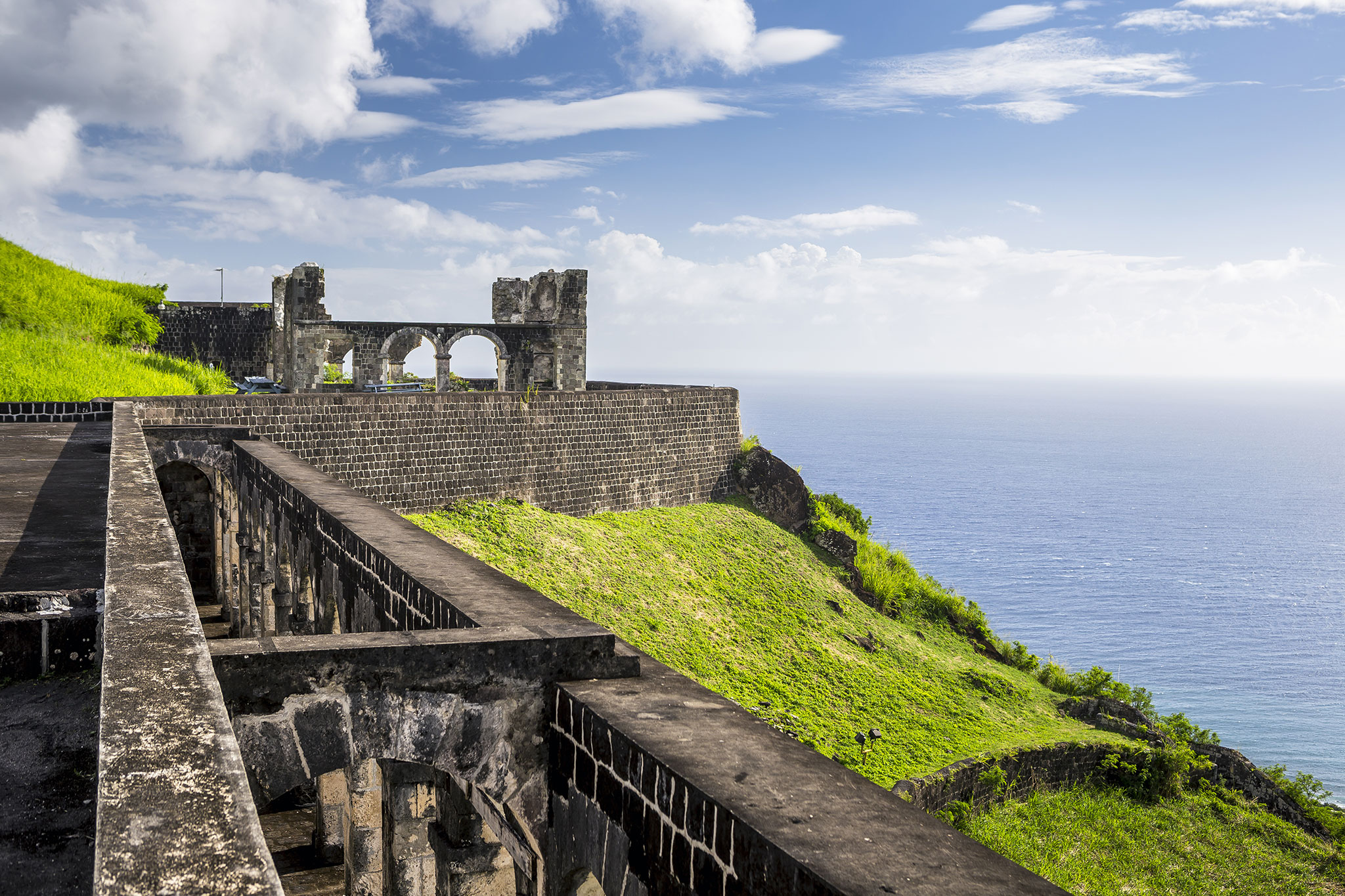 Ancient fortress ruins on a lush green cliff overlooking the sea, with archways and stone walls, under a blue sky with scattered clouds.