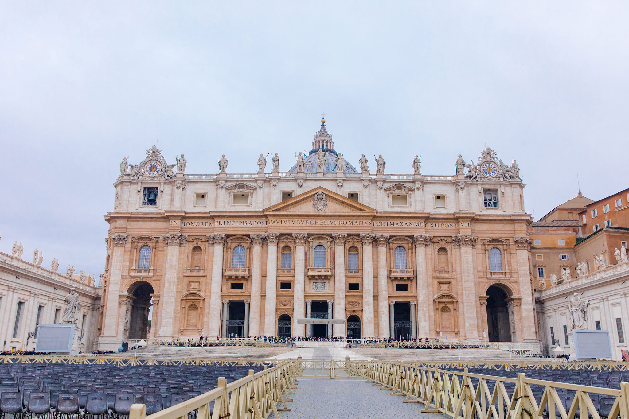 The facade of St. Peter’s Basilica in Vatican City, showcasing its detailed Renaissance architecture and rows of empty seating in the foreground, under an overcast sky.