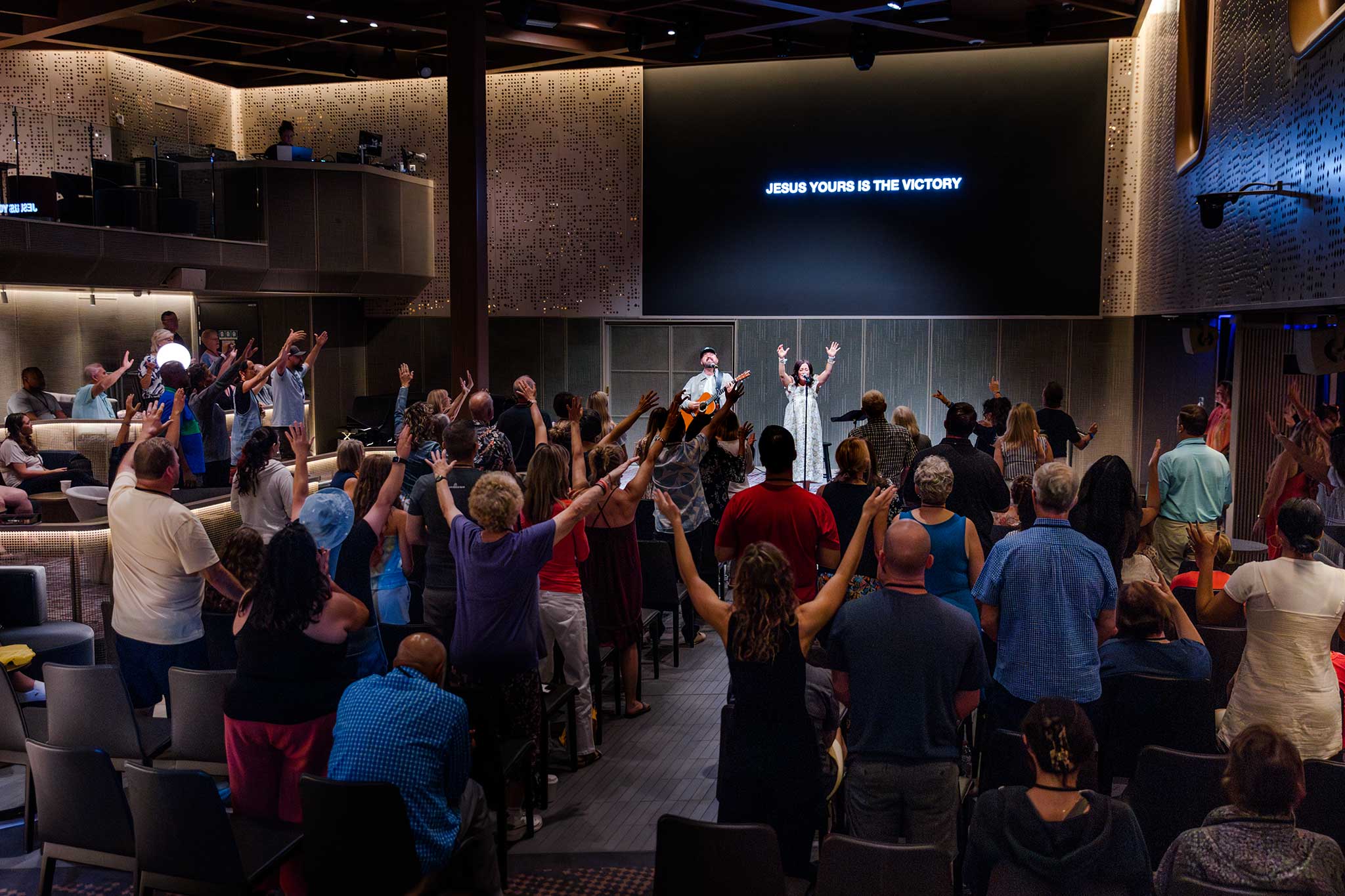 Congregation with raised hands engaging in worship led by a guitarist and singer on stage.