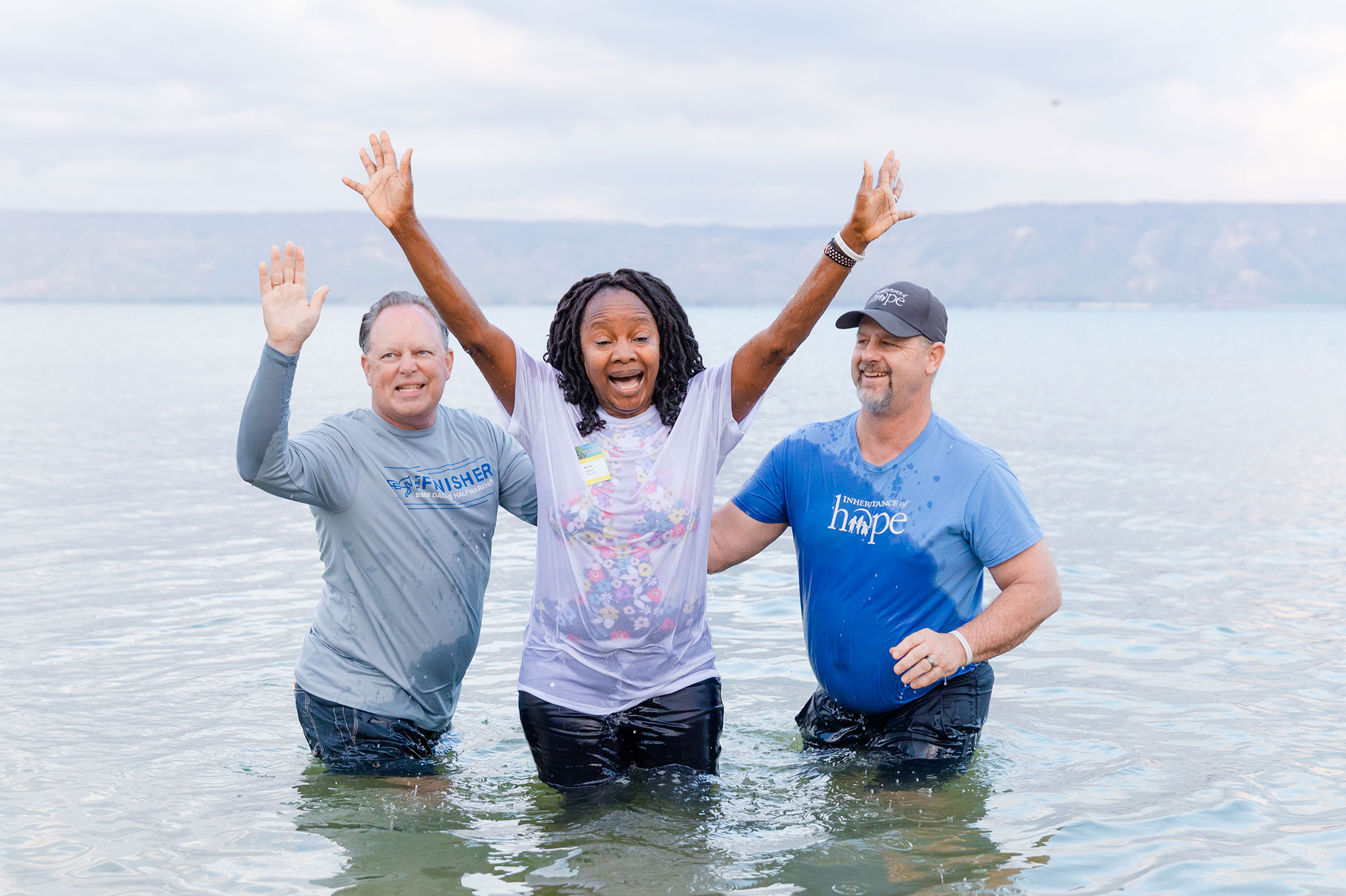 Three joyful adults raise their hands in joy while standing in shallow water, with a hilly landscape in the background.