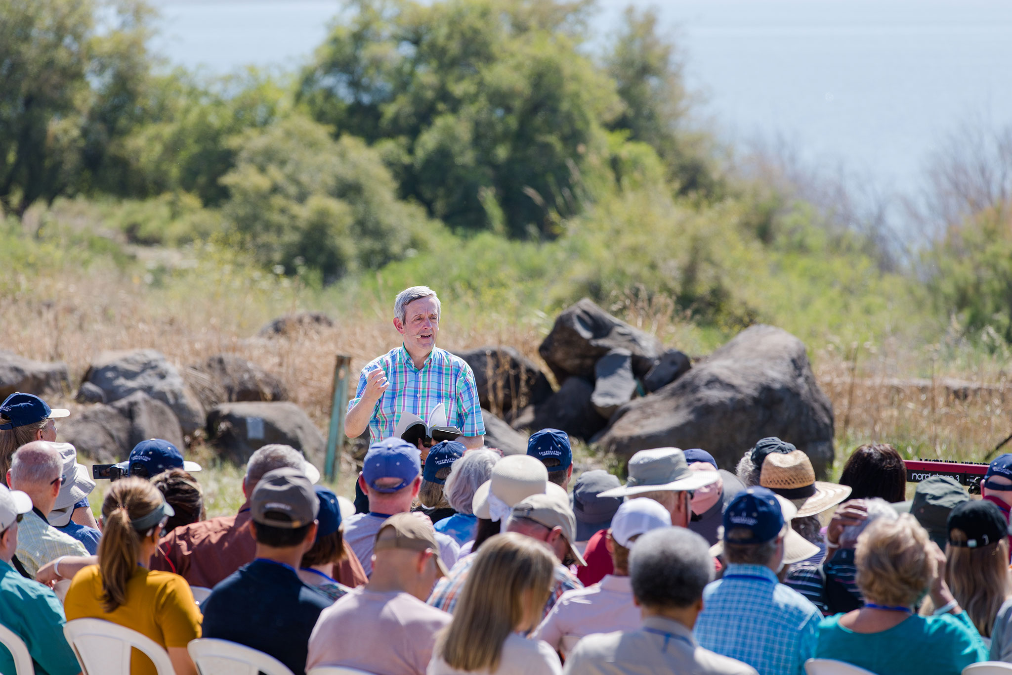 A man lecturing outdoors to a seated crowd of diverse people with a scenic backdrop of water and greenery.