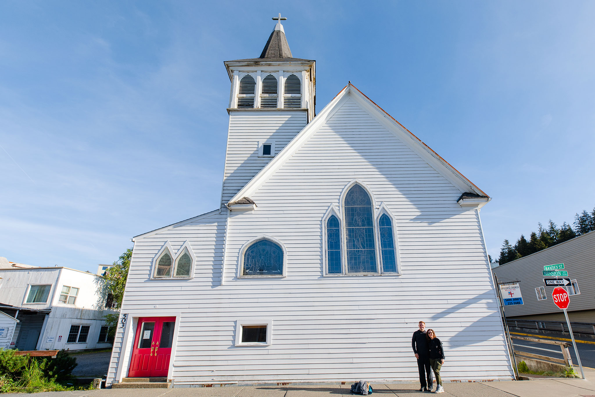 Two people posing in front of a traditional white church with stained glass windows, a red door and a bell tower under a blue sky.