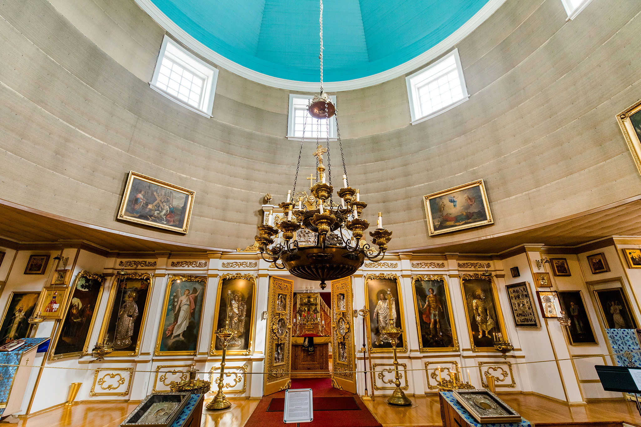 Ornate church interior with a striking blue dome, large central chandelier and walls covered in religious artwork and icons.