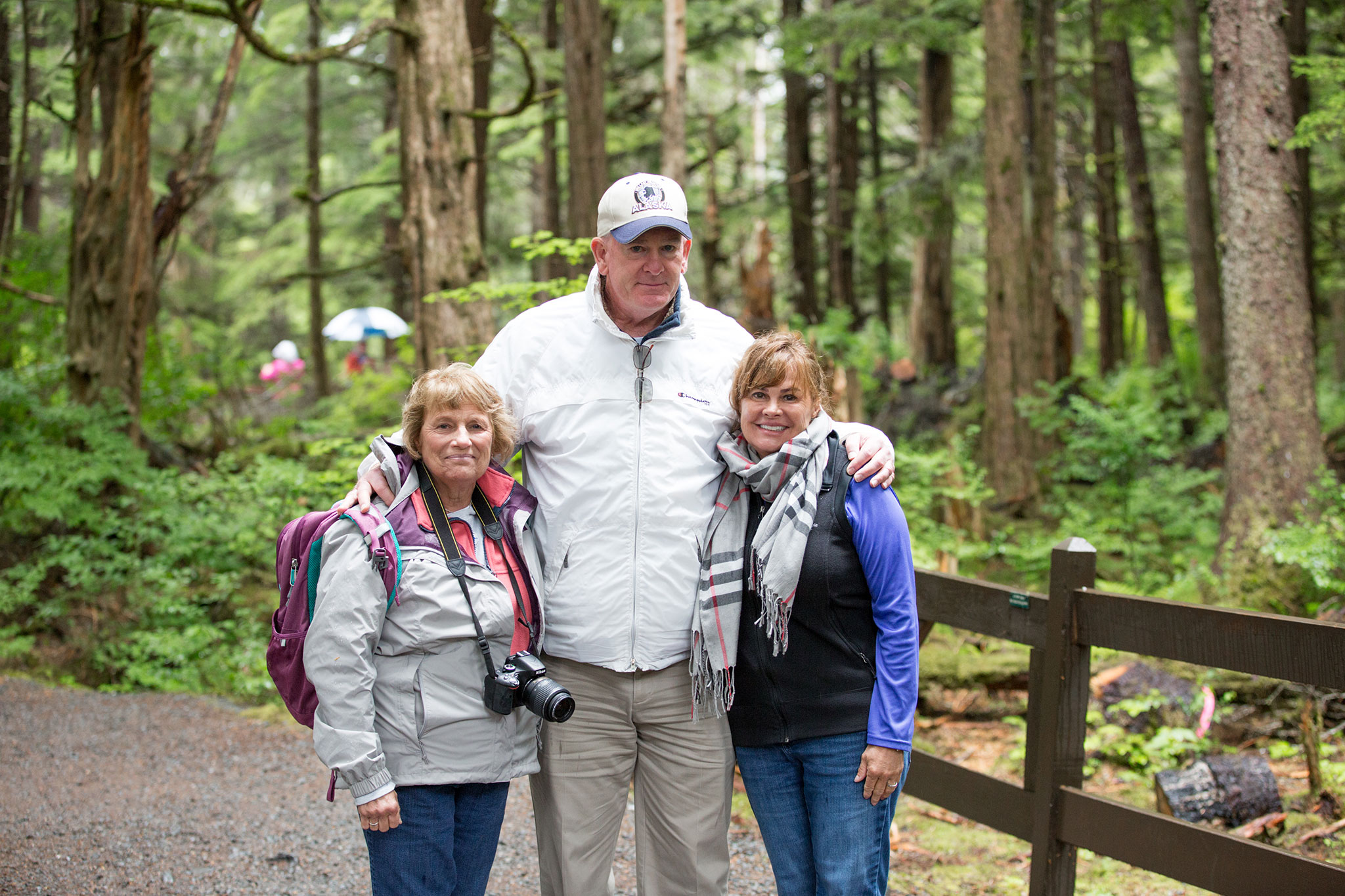 A group of three hikers, two women, and one man, smiling in a wooded area with tall trees in the background.