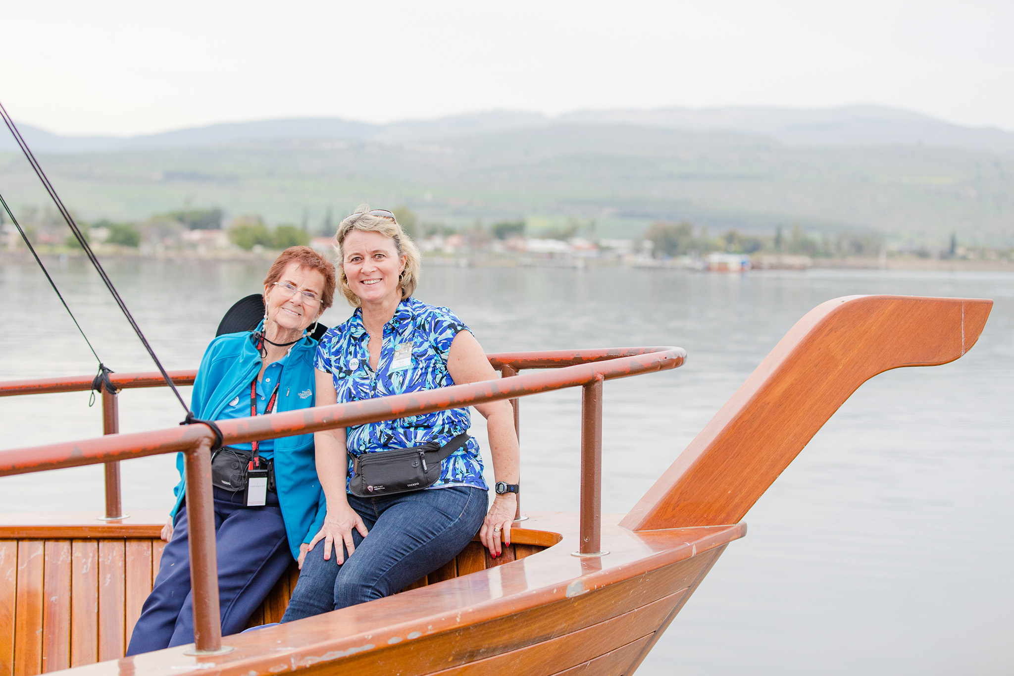 Two smiling ladies with lanyards sitting on a wooden boat, scenic hills and water behind them.