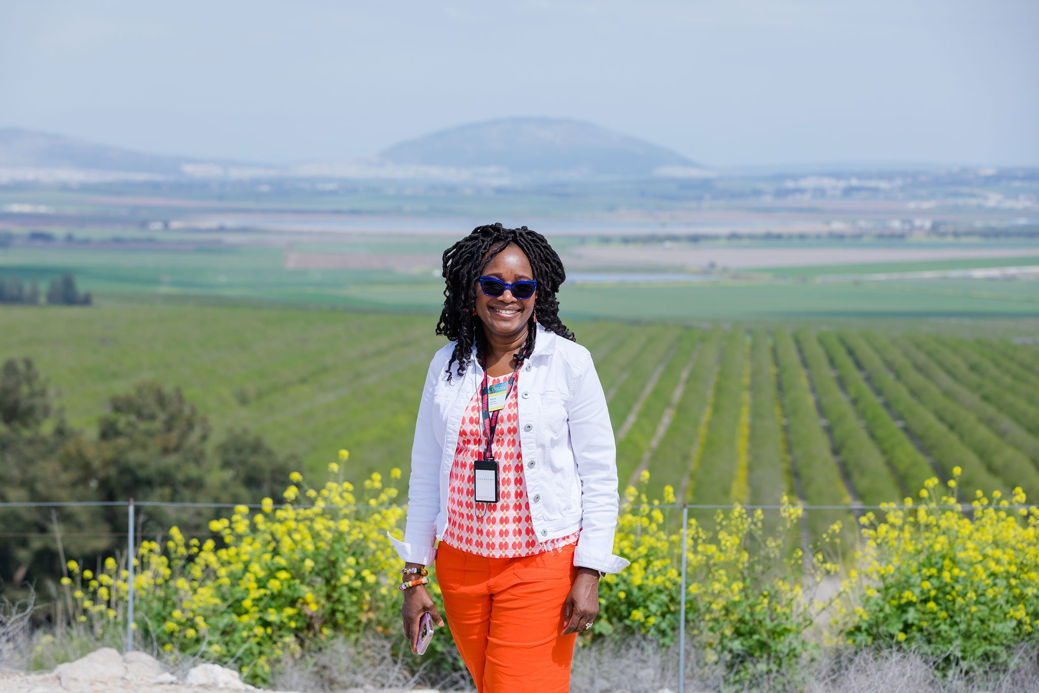 A happy traveler in bright colors poses in front of a lush rural landscape with vineyards and yellow wildflowers under a clear sky.