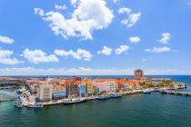 Panoramic view of a bustling Caribbean harbor in Curacao, featuring vibrant, multicolored buildings and a busy waterfront with boats and clear blue skies.