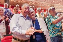 An elderly couple embracing and smiling joyfully at a social event, surrounded by a crowd of applauding attendees.