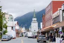 A bustling street scene in a small town with a white clock tower in the center, surrounded by mountains and colorful storefronts.