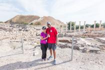 A couple smiling at the camera, standing in front of ancient ruins with tall columns and a hill in the background.