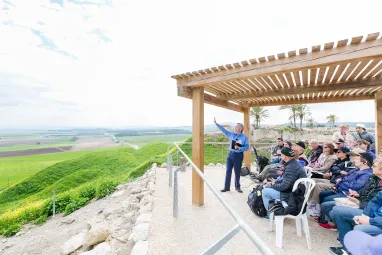 Dr. Robert Jeffress holding a book and gesturing toward a panoramic view of lush hills and fields, with tourists seated under a pergola listening intently.