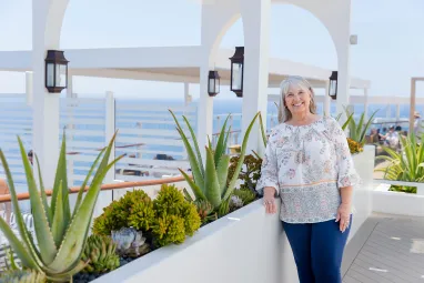 A cheerful senior woman with grey hair, smiling and standing on a cruise ship balcony with ocean views and decorative plants.