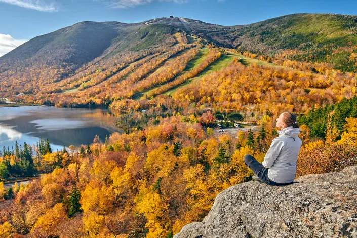 Autumn view of a colorful forest and lake in New England, with a woman in a white jacket seated peacefully on a large rock, enjoying the panoramic scenery.